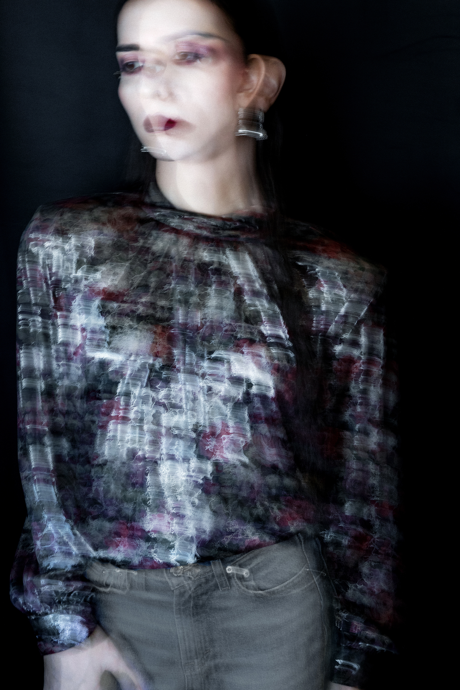 Blurry image of a female model with pale skin and deep blush in a foiled blouse against a dark backdrop